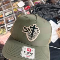 Olive trucker hat with a Rooted in Christ patch showing a cross with roots